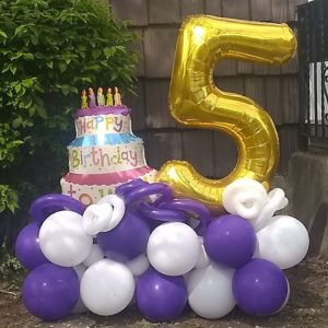 Birthday celebration balloons with a large golden number 5 foil balloon and a foil birthday cake balloon over round purple and white balloons